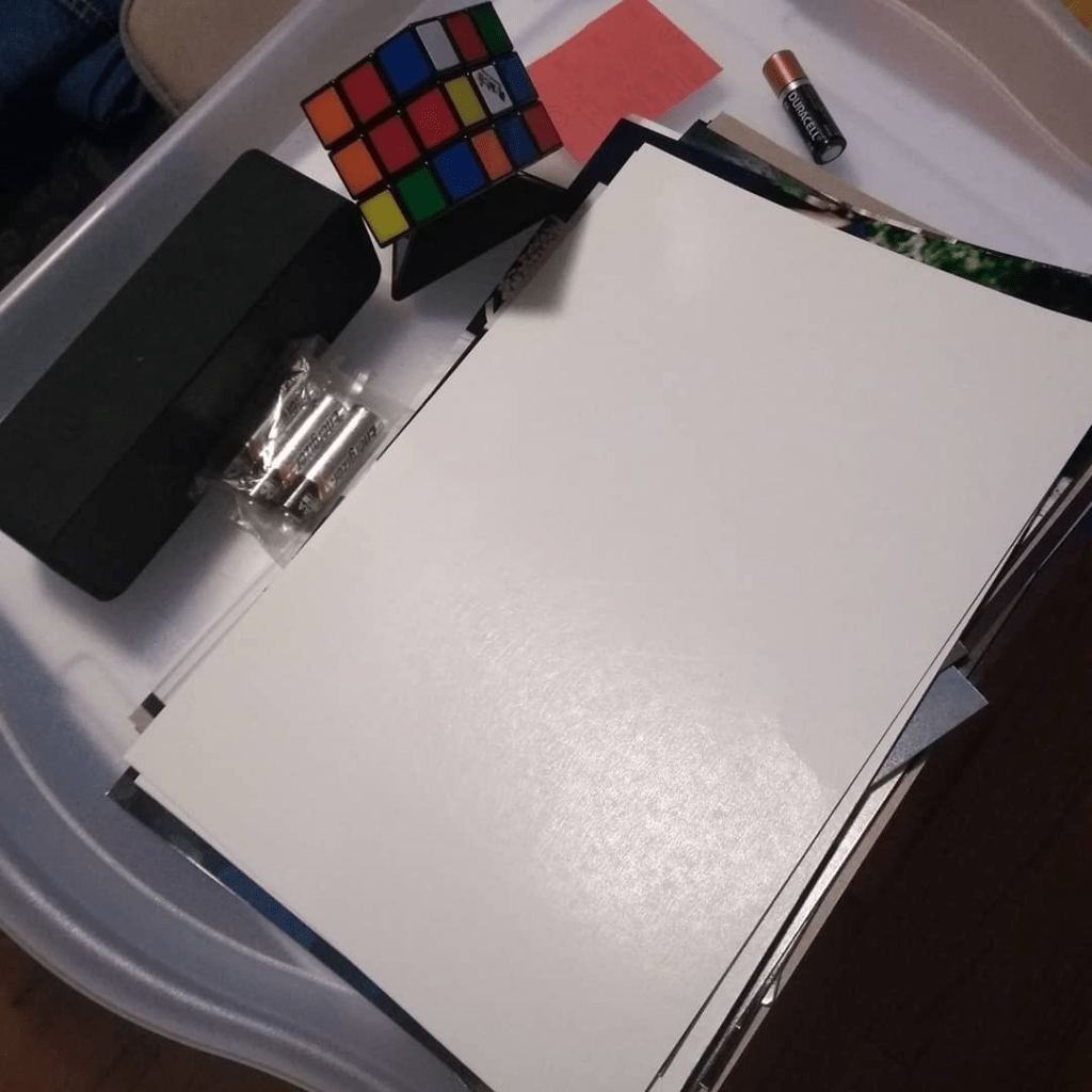 A tray with various items taken off of a bookshelf because they didn’t seem to belong on the bookshelf. The items include: loose photographs, a speaker, batteries, and a Rubik’s cube. This image is a part of the project “A retrospective on decluttering books” by Aden Hailemariam on adenhailemariam.com.